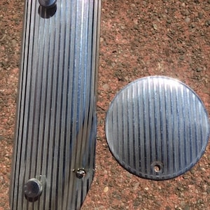 May include: Two silver-coloured metal gun grips with a grooved texture. The grips have a small round knob on the top and a small hole on the bottom.