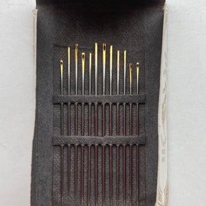 Op de afbeelding: Een doos met verschillende naalden, met een zwarte viltvoering en een wit label met gouden letters waarop "The Best Needles Made" staat.