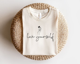 May include: A beige t-shirt with a black graphic of a hand holding hearts and the text "love yourself" in black script.