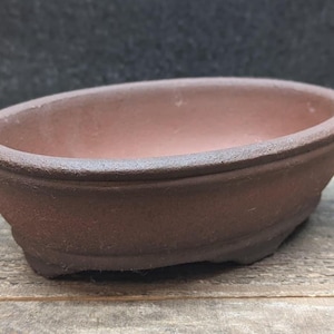 May include: An oval-shaped, unglazed brown ceramic bonsai pot with a textured, matte finish. The vessel features a thick, rounded rim, a deep basin, and small, integrated feet at the base, providing a stable foundation for small plant displays.