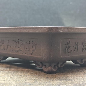 May include: A rectangular brown bonsai pot with a matte finish, featuring four decorative feet and embossed Chinese characters on the side that read 花开富贵, alongside intricate relief carvings of scrolling vines and floral patterns.