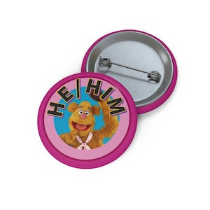 Op de afbeelding: Roze knoop met een afbeelding van een bruine beer op een blauwe achtergrond en de tekst "HE/HIM" in zwarte en gele letters.