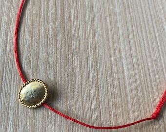 Könnte beinhalten: Ein goldfarbener ovaler Anhänger mit einem seilartigen Rand ist an einer dünnen roten Schnur aufgefädelt. Die Schnur ist auf beiden Seiten des Anhängers verknotet. Der Hintergrund ist eine hellfarbige Holzoberfläche.