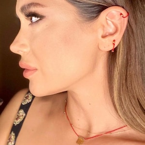 Puede incluir: Una mujer lleva un collar de cuerda roja con un colgante de oro. Lleva un pendiente de cuerda roja en la oreja izquierda y un pendiente de aro de oro en la oreja derecha.