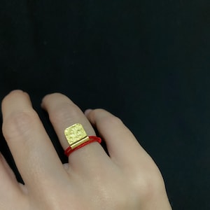 Op de afbeelding: Een gouden ring met een getextureerd vierkant ontwerp, op een rood touwtje.