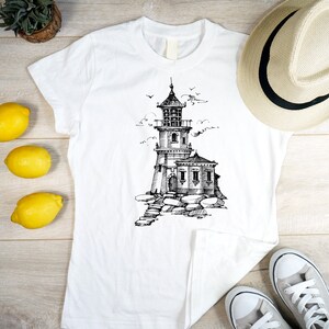 Puede incluir: Camiseta blanca con una ilustraci&oacute;n en blanco y negro de un faro en una costa rocosa. La camiseta est&aacute; colocada sobre una superficie de madera con un sombrero de paja, limones y zapatillas deportivas.