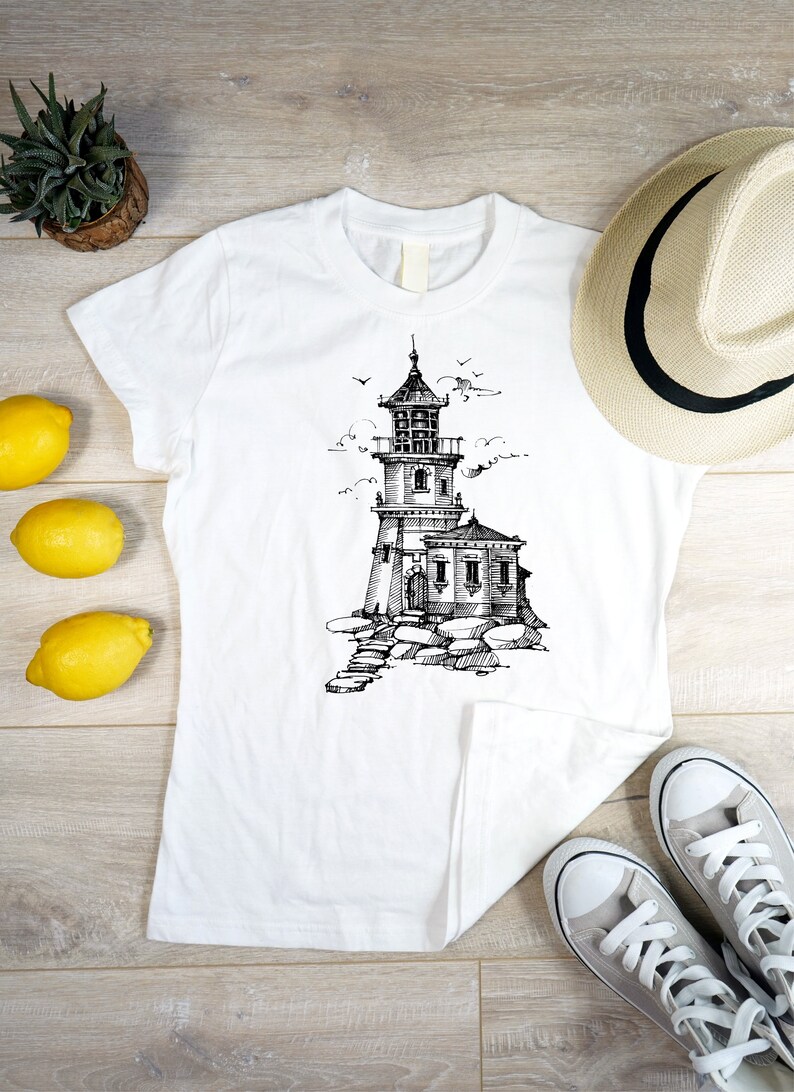 Puede incluir: Camiseta blanca con una ilustraci&oacute;n en blanco y negro de un faro en una costa rocosa. La camiseta est&aacute; colocada sobre una superficie de madera con un sombrero de paja, limones y zapatillas deportivas.
