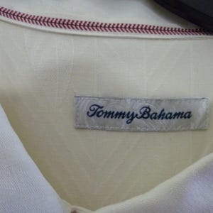Puede incluir: Primer plano de una camisa color crema con la etiqueta "Tommy Bahama" visible. La camisa tiene un patr&oacute;n texturizado y un detalle cosido en rojo a lo largo del cuello. La etiqueta es rectangular con el nombre de la marca en azul oscuro.