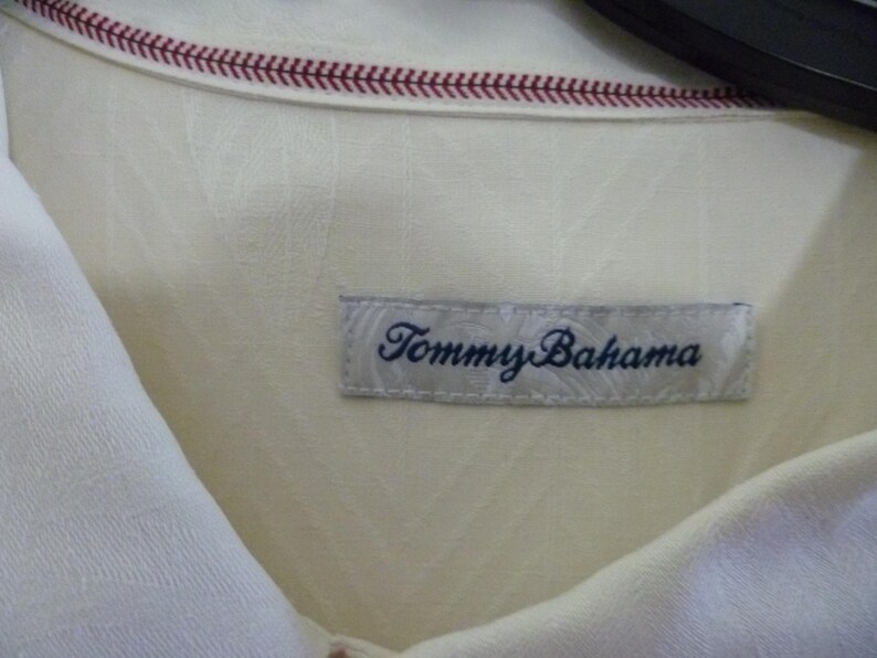 Puede incluir: Primer plano de una camisa color crema con la etiqueta "Tommy Bahama" visible. La camisa tiene un patr&oacute;n texturizado y un detalle cosido en rojo a lo largo del cuello. La etiqueta es rectangular con el nombre de la marca en azul oscuro.