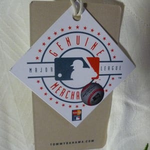 Puede incluir: Una etiqueta en forma de diamante con las palabras "GENUINE MERCHANDISE" que rodean un logotipo de la Major League Baseball. Se adjunta una pegatina de b&eacute;isbol m&aacute;s peque&ntilde;a. La etiqueta es de color canela con una cuerda blanca y el sitio web TOMMYBAHAMA.COM.