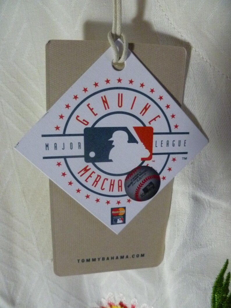 Puede incluir: Una etiqueta en forma de diamante con las palabras "GENUINE MERCHANDISE" que rodean un logotipo de la Major League Baseball. Se adjunta una pegatina de b&eacute;isbol m&aacute;s peque&ntilde;a. La etiqueta es de color canela con una cuerda blanca y el sitio web TOMMYBAHAMA.COM.