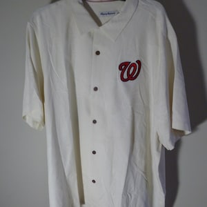 Puede incluir: Camisa crema con botones y mangas cortas. Presenta un logotipo "W" rojo y azul marino en la parte delantera. La camisa tiene botones marrones oscuros y un cuello. La camisa est&aacute; colgada de una percha negra.