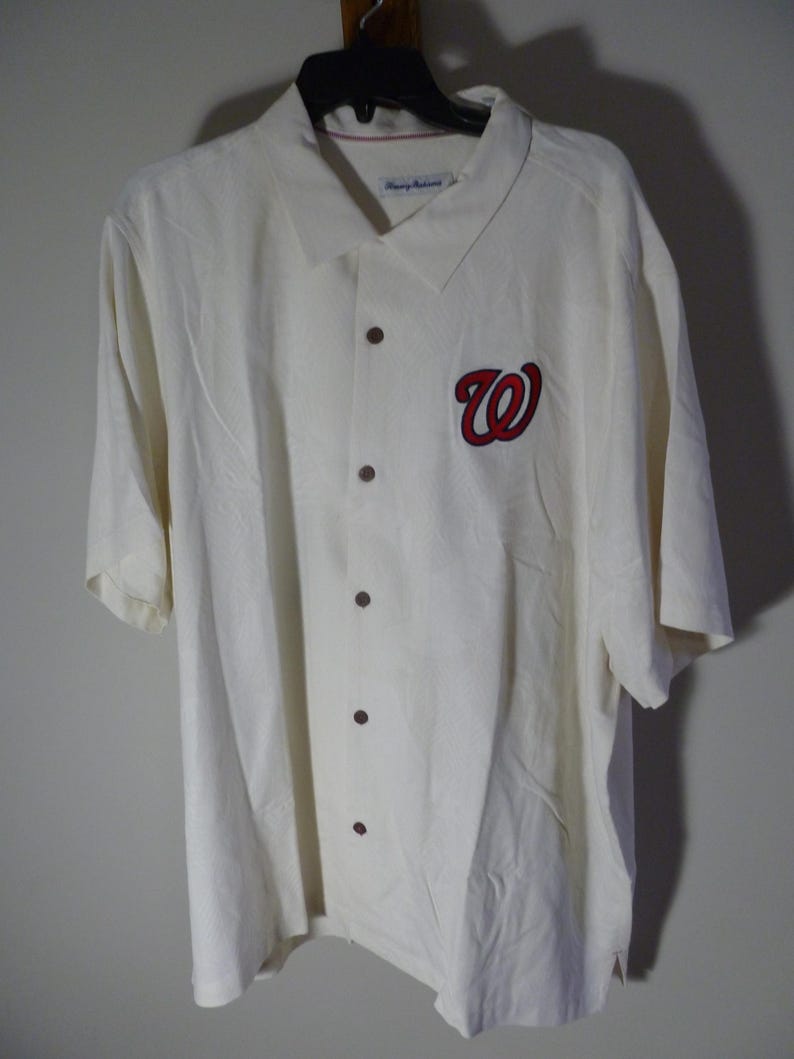 Puede incluir: Camisa crema con botones y mangas cortas. Presenta un logotipo "W" rojo y azul marino en la parte delantera. La camisa tiene botones marrones oscuros y un cuello. La camisa est&aacute; colgada de una percha negra.