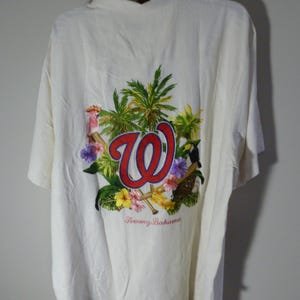 Puede incluir: Camisa crema de manga corta con dise&ntilde;o tropical. La parte trasera presenta un gran gr&aacute;fico con las letras "W" en rojo, rodeadas de palmeras, flores y p&aacute;jaros. La marca "Tommy Bahama" est&aacute; impresa debajo.