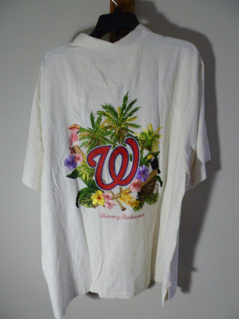 Puede incluir: Camisa crema de manga corta con dise&ntilde;o tropical. La parte trasera presenta un gran gr&aacute;fico con las letras "W" en rojo, rodeadas de palmeras, flores y p&aacute;jaros. La marca "Tommy Bahama" est&aacute; impresa debajo.