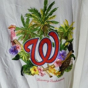 Puede incluir: Camisa blanca con botones con un dise&ntilde;o bordado con un logotipo "W" rojo y azul, palmeras, flores y un tuc&aacute;n. La camisa tambi&eacute;n tiene el texto bordado "Tommy Bahama".