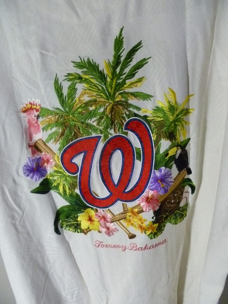 Puede incluir: Camisa blanca con botones con un dise&ntilde;o bordado con un logotipo "W" rojo y azul, palmeras, flores y un tuc&aacute;n. La camisa tambi&eacute;n tiene el texto bordado "Tommy Bahama".