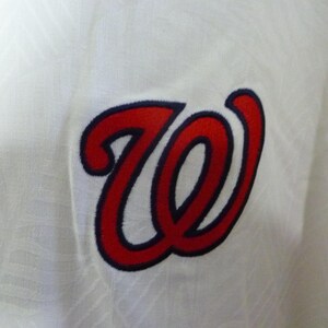 Puede incluir: Primer plano de una tela blanca con un logotipo "W" bordado en rojo y azul marino. El logotipo es una letra estilizada con un contorno en negrita, probablemente para un equipo deportivo o una marca.