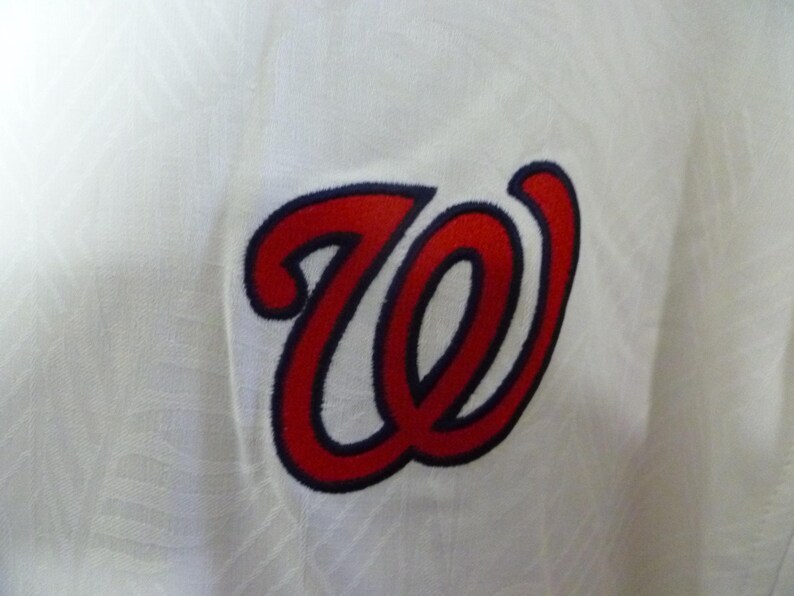 Puede incluir: Primer plano de una tela blanca con un logotipo "W" bordado en rojo y azul marino. El logotipo es una letra estilizada con un contorno en negrita, probablemente para un equipo deportivo o una marca.