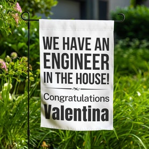 May include: A white garden flag with black text that reads "WE HAVE AN ENGINEER IN THE HOUSE!" and "Congratulations Valentina." The flag is displayed in a garden with green foliage and pink flowers.
