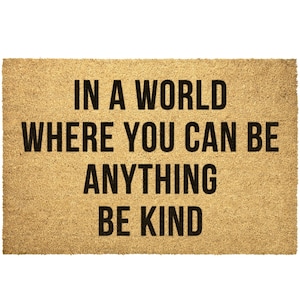 May include: A brown doormat with the text "In a world where you can be anything be kind" in black lettering.