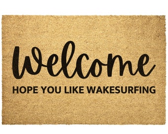 May include: A brown coir doormat with the text "welcome" in black cursive lettering and the text "HOPE YOU LIKE WAKESURFING" in black block lettering.
