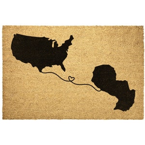 May include: A brown coir doormat with a black silhouette of the United States and a black silhouette of a smaller landmass connected by a line with a heart in the middle.