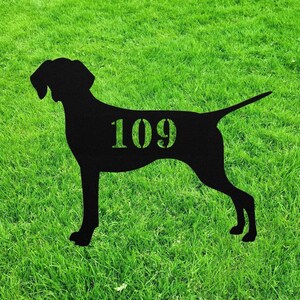 May include: A black metal silhouette of a dog, standing on green grass. The number "109" is cut out of the dog's body. The dog has a long tail and pointed ears. The grass is a vibrant green.