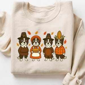 May include: Beige sweatshirt featuring a Thanksgiving-themed graphic. The graphic depicts four cartoon dogs dressed in pilgrim and harvest attire, surrounded by autumn leaves. The dogs are wearing hats, aprons, and a corn husk.