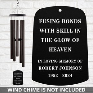May include: A black metal memorial plaque with the text "FUSING BONDS WITH SKILL IN THE GLOW OF HEAVEN" and "IN LOVING MEMORY OF ROBERT JOHNSON 1952-2024". A wind chime with dark brown tubes hangs to the left.