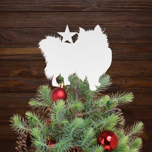 May include: A Christmas tree with red ornaments and pine cones. A white silhouette of a dog with a star on top is placed on the tree. The background is a dark brown wooden surface.
