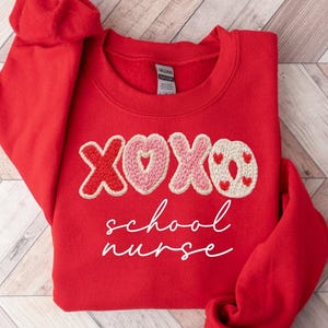 May include: A red sweatshirt with the embroidered letters "XOXO" in red and pink, with a heart in the center of the "O". The words "school nurse" are written in white script below the letters. The sweatshirt has long sleeves.