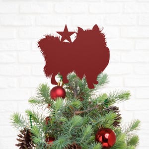 May include: A burgundy-colored dog silhouette with a star accent, placed atop a Christmas tree. The tree is decorated with red ornaments and pine cones. The background is a white brick wall.