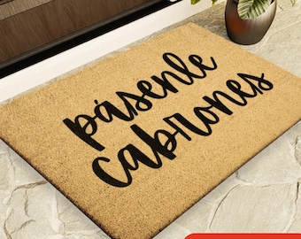 May include: A rectangular doormat made of natural fibers, featuring the phrase "Pasenle Cabrones" in bold, black script. The mat is tan with a dark brown border. The bottom right corner has a red circle with the text "4 SIZES".