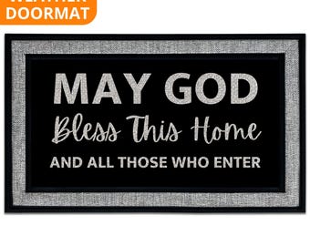 May include: Black rectangular all-weather doormat with a silver border. The mat features the text "MAY GOD Bless This Home AND ALL THOSE WHO ENTER" in white, stylized lettering. The top left corner has the text "ALL WEATHER DOORMAT" in orange.