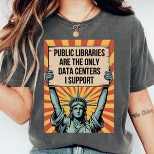 May include: A charcoal gray t-shirt featuring a graphic of the Statue of Liberty holding a sign that reads, "PUBLIC LIBRARIES ARE THE ONLY DATA CENTERS I SUPPORT." The background of the graphic consists of a yellow and red sunburst pattern.
