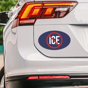 May include: An oval sticker with a navy blue background and the word "ICE" in white, crossed out by a red circle. The sticker is affixed to the rear of a white vehicle.