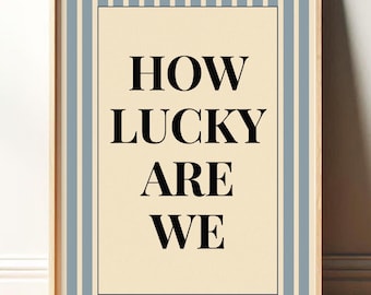 May include: A framed art print with the text "HOW LUCKY ARE WE" in black, centered on a cream-colored background. The print is bordered by a black rectangle and framed with light blue vertical stripes. The frame is made of light wood.