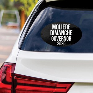 May include: An oval-shaped black car decal applied to the rear window of a white vehicle. The sticker features white sans-serif text arranged in four lines that reads: MOLIERE, DIMANCHE, GOVERNOR, 2026. The design is centered on the glass surface.