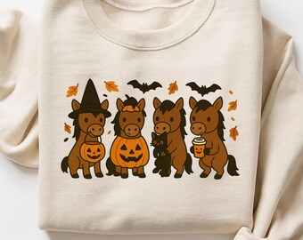 May include: A beige sweatshirt featuring a Halloween-themed design. The graphic shows four brown horses dressed in costumes, including a witch hat, pumpkin, and holding a coffee cup. Black bats and orange leaves complete the festive design.