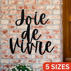 Puede incluir: Decoraci&oacute;n de pared de metal negro con la frase francesa "Joie de Vivre" en una fuente cursiva. La decoraci&oacute;n est&aacute; montada en una pared de ladrillo. El texto "5 TAMA&Ntilde;OS" es visible en la esquina inferior derecha de la imagen.