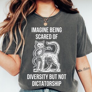 May include: Dark gray t-shirt with the text "IMAGINE BEING SCARED OF DIVERSITY BUT NOT DICTATORSHIP". The graphic shows a cat with a snake in its mouth. The shirt is a casual, unisex style.