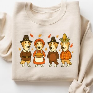 May include: Beige sweatshirt featuring a Thanksgiving-themed illustration. Four golden retriever dogs are dressed in festive attire, including pilgrim hats, a bonnet, and a corn husk hat. Autumn leaves are scattered around the dogs.