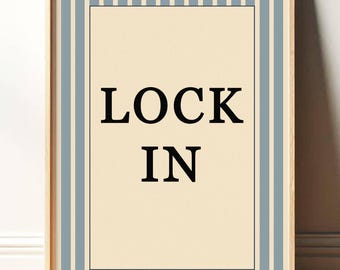 May include: Framed wall art with the words "LOCK IN" in bold black letters. The artwork has a cream-colored background with a black border. Vertical light blue stripes frame the artwork, and the frame is made of light wood.