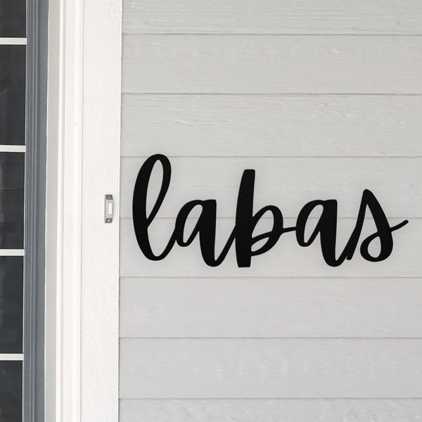 May include: Black metal sign with the word "labas" written in a cursive font. The sign is mounted on a white wooden wall.