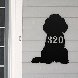 May include: Black metal silhouette of a dog sitting with the house number 320 on its back. The silhouette is mounted on a gray wooden wall.