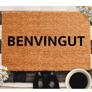 May include: A rectangular doormat with the word "BENVINGUT" in black letters. The mat is brown and sits on a black and white checkered pattern. White daisies and a cup of coffee are in the image.