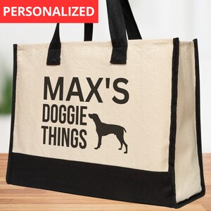 May include: A beige canvas tote bag with black handles and trim. The bag has the text "MAX'S DOGGIE THINGS" printed on it in black, along with a black silhouette of a dog.
