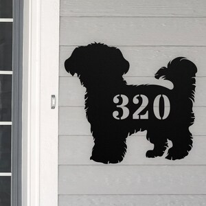 May include: Black metal silhouette of a dog with the house number 320 cut out of the silhouette. The silhouette is mounted on a gray wooden surface.