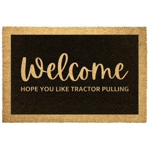 May include: A brown coir doormat with a black border and the text "Welcome" in gold script. The text "Hope you like tractor pulling" is written in gold below the welcome message.